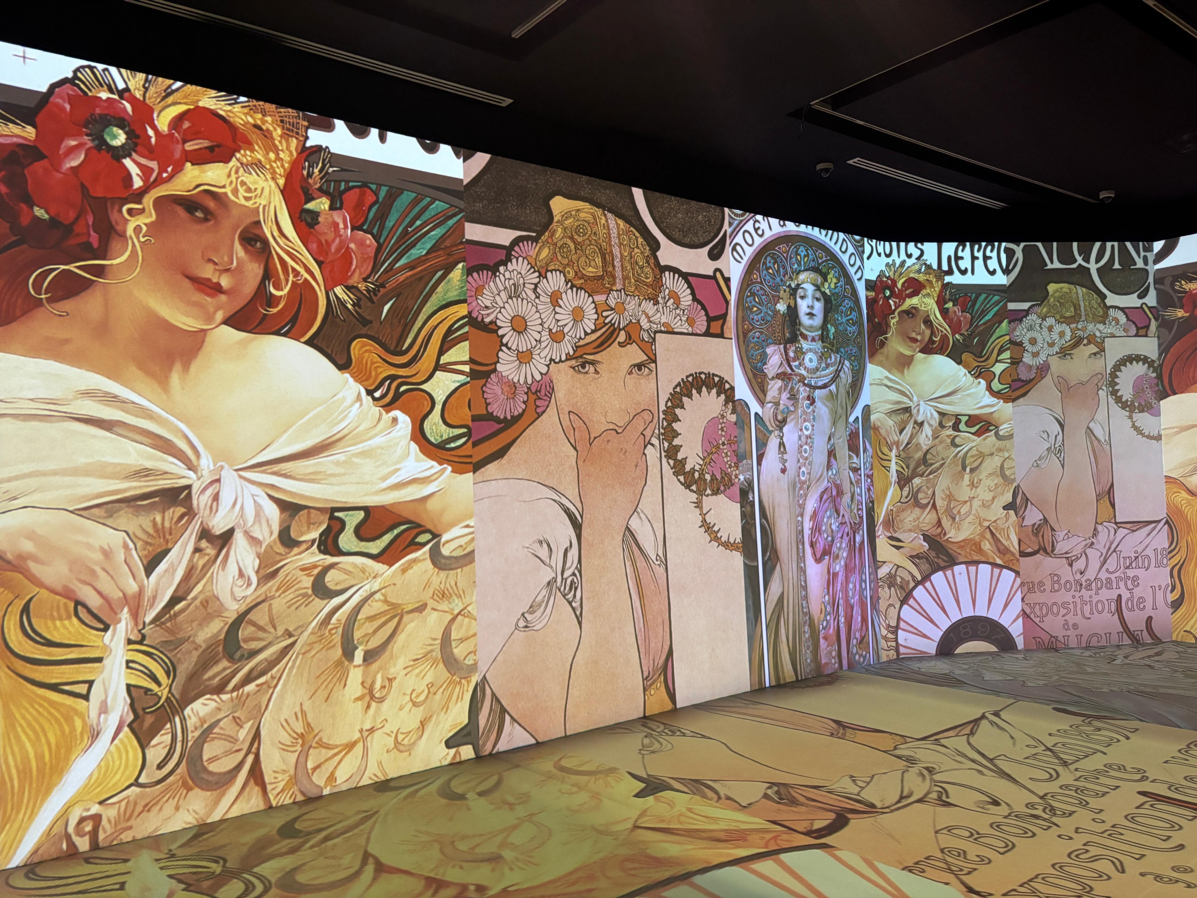 Grandes pantallas iluminadas en una sala oscura proyectan ilustraciones al estilo de Alphonse Mucha con retratos de mujeres estilizadas