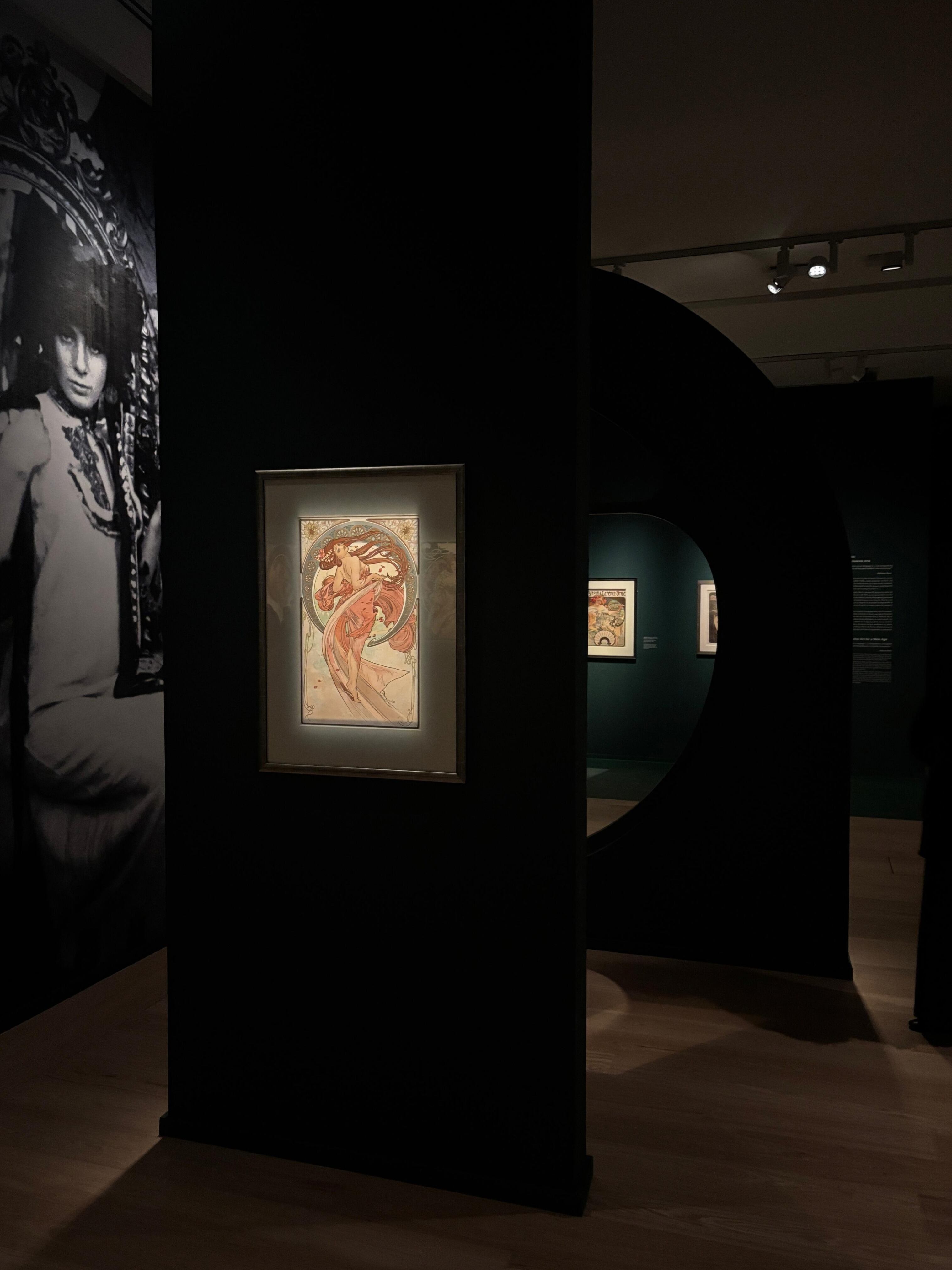 Un cuadro ilustrado colgado en una pared negra junto a una fotografía en blanco y negro y un panel con apertura circular